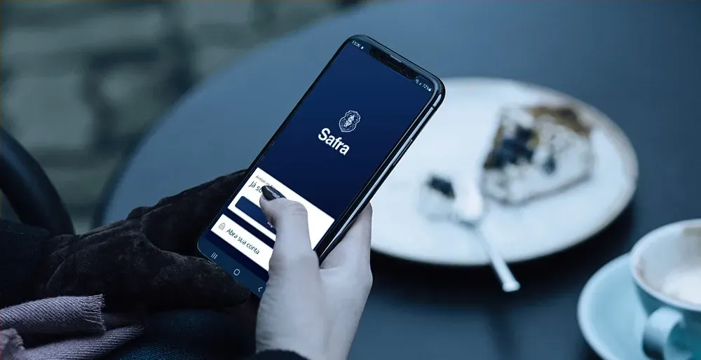 A person's hand using a smartphone, showing the Stake app, next to a plate with a dessert and a coffee cup.