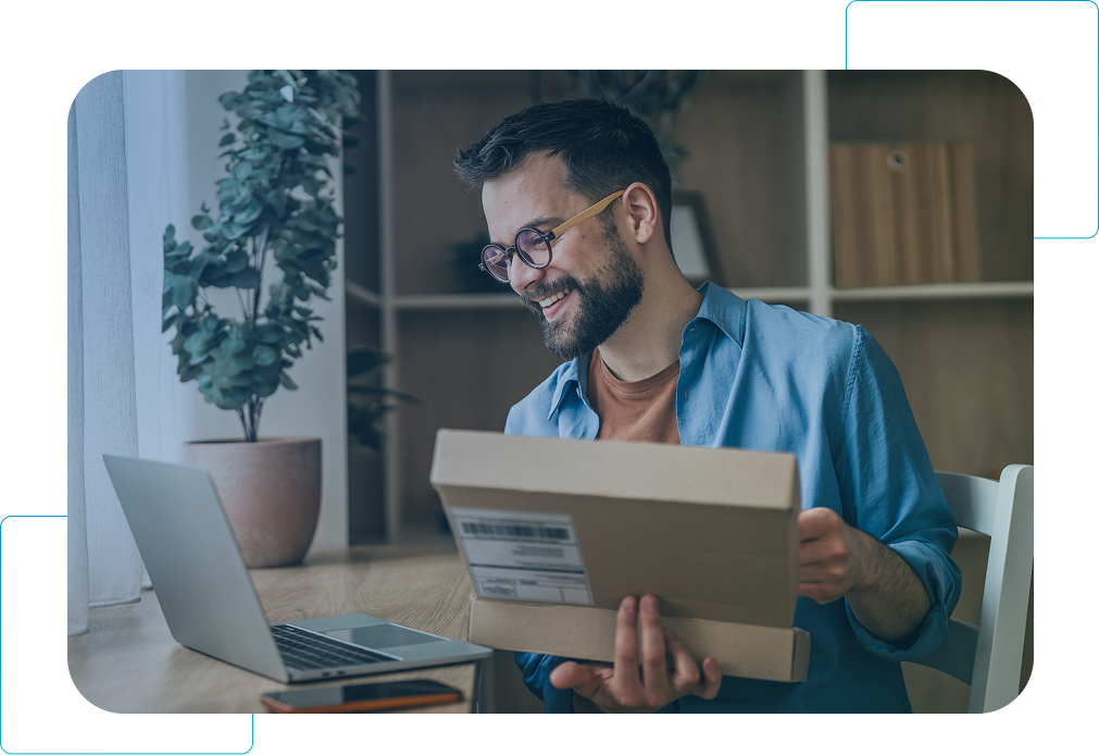 Man smiling while holding a box and checking a laptop at a desk in a bright home office