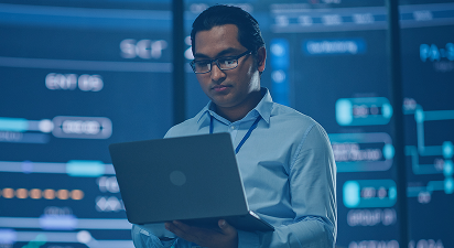 Man in glasses holding a laptop, standing in front of illuminated screens in a tech setting.