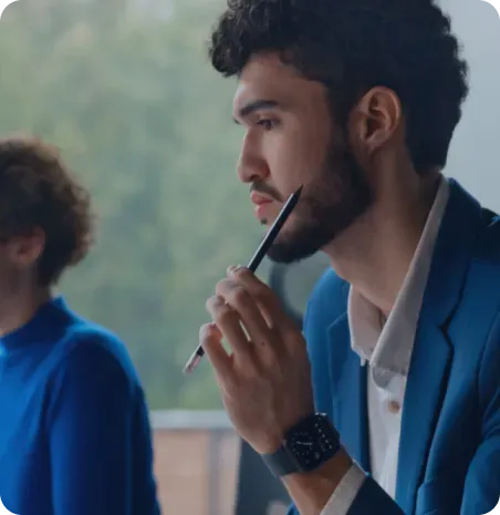 Man in blue blazer contemplates, holding a pencil near his mouth; another person partially visible in blue.