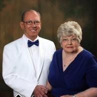 Two people posed against a dark backdrop, one in a white tuxedo and blue bow tie, the other in a navy dress.