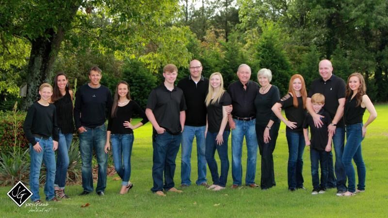 Group of people standing on grass in a park, wearing mostly black tops and blue jeans, with trees behind them
