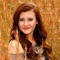 Woman with long brown hair and red lipstick standing in tall golden grass outdoors.
