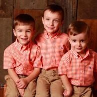 Three children in matching red checkered shirts pose against a wood-paneled backdrop.
