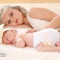 Sleeping baby in a white diaper beside a blonde child lying on a soft white blanket