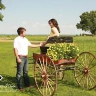 Man standing by a woman seated in a flower-filled cart on a grassy field under a bright sky