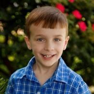 Smiling child in a blue plaid shirt outdoors with blurred greenery and red flowers in the background
