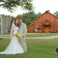 Bride and groom posing on a lawn in front of a wooden barn, holding a bouquet.
