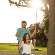A man standing behind a woman sitting on a swing at sunset in a grassy yard, with trees and a house in the background.