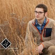 Person sitting in tall dry grass wearing glasses and a tan jacket, looking to the right