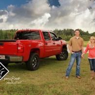Red pickup truck in a field with two people standing beside it under a cloudy sky