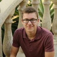 Smiling man in glasses and maroon polo sitting on cement stone steps with columns in the background outdoors
