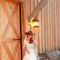 Woman in a white dress and brown cowboy hat standing by a wooden barn door and lantern