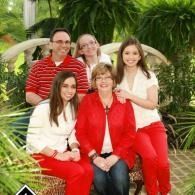 Five people posing together in a lush green garden, wearing red, white, and a red jacket.