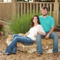 Couple sitting on a large rock outdoors in front of a wooden fence, smiling at the camera.