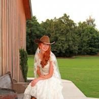 Woman in a white dress and brown cowboy hat sitting outdoors beside a wooden building, with a green lawn behind her.