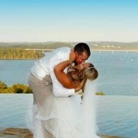 Couple embracing by a calm lakeside at sunset, with blue water and distant shoreline.