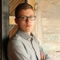 Portrait of a person in glasses and a gray shirt standing by a wooden wall
