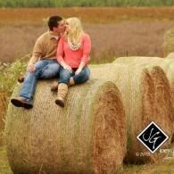 Couple sitting on a hay bale in a field, facing each other and smiling.