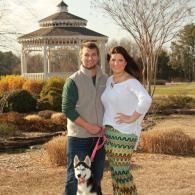 Couple standing with a dog in a park near a gazebo, with winter trees and shrubs behind them.