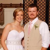 Couple in wedding attire posing indoors, with the bride in a white dress and the groom in a beige vest.