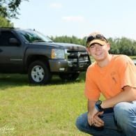 Man crouching on grass beside a dark pickup truck under a bright sky