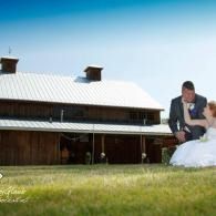 Bride and groom seated on grass in front of a rustic barn under a bright sky