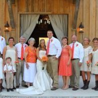 Wedding party posing outside a rustic wooden venue, with the bride and groom centered amid family and friends.