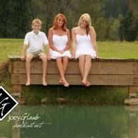 Three children in white sit on a wooden bridge above a green pond, with trees in the background.