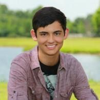 Smiling person in a pink shirt outdoors by a lake with green trees in the background