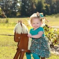Smiling toddler in teal outfit riding a wooden toy horse outdoors in a sunny field