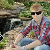 Person in sunglasses and a red plaid shirt sitting by a small waterfall in a rocky garden.