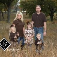 Family of four posing outdoors in a grassy field, with trees in the background.