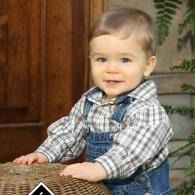 Smiling baby in blue overalls and plaid shirt, leaning on a woven basket indoors.