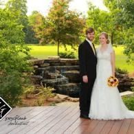 Bride and groom pose on a wooden deck beside a small waterfall and lush green garden.