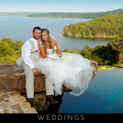 Wedding couple in white on a rocky ledge by a blue lake with a forested shoreline and pool below