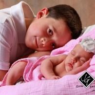 A child gently leans over a sleeping baby on a pink blanket.