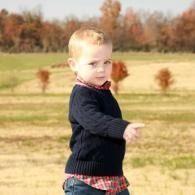 Young child standing outdoors in a field, wearing a dark sweater and plaid shirt, pointing to the side.