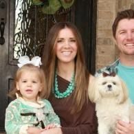 Woman holding a child and a small white dog on a front porch, all smiling