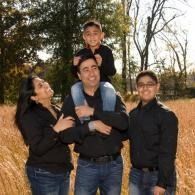 Family of four standing outdoors in a grassy field, smiling with a child on a man's shoulders.