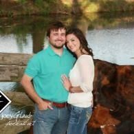Couple posing by a lake, smiling and hugging, with a blanket hanging on a wooden fence in the background