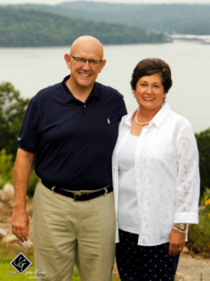 Two smiling people standing outdoors by a lake, with trees and hills in the background.