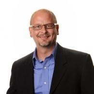 Smiling man in glasses wearing a black blazer and blue shirt against a white background