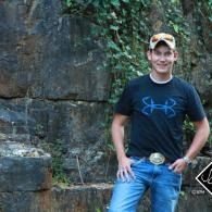 Man standing by a rocky cliff with vines, wearing a black T-shirt, jeans, and a cap, smiling at the camera