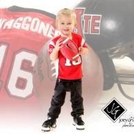 Child in a red sports jersey and black pants standing in front of a football backdrop and holding a toy football