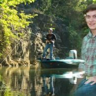 Man on a boat in a narrow, tree-lined river, with another person sitting on the right foreground.