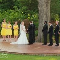 Wedding ceremony on a wooden deck, bride and groom facing each other with attendants standing nearby outdoors.