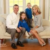 Family of four posing on a beige couch in a bright living room, all dressed in blue and neutral tones.
