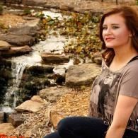Woman sitting by a rocky stream with a small waterfall, wearing a gray shirt and dark jeans.