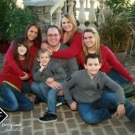 Family posing together outdoors on a stone path, dressed in red and gray sweaters, smiling at the camera.